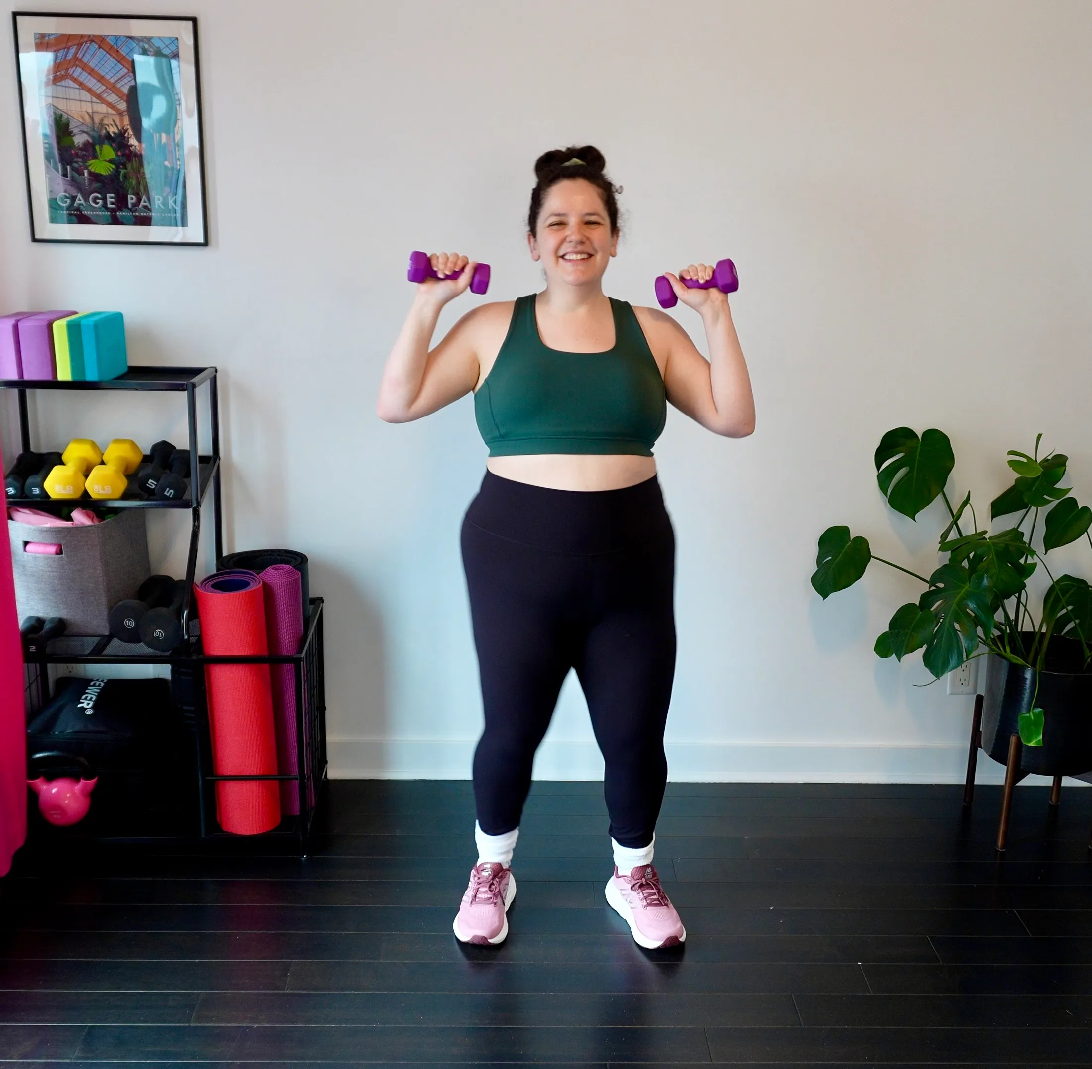 Rebecca Yaffa smiling in her home gym, holding purple dumbbells at her shoulders in workout clothes.